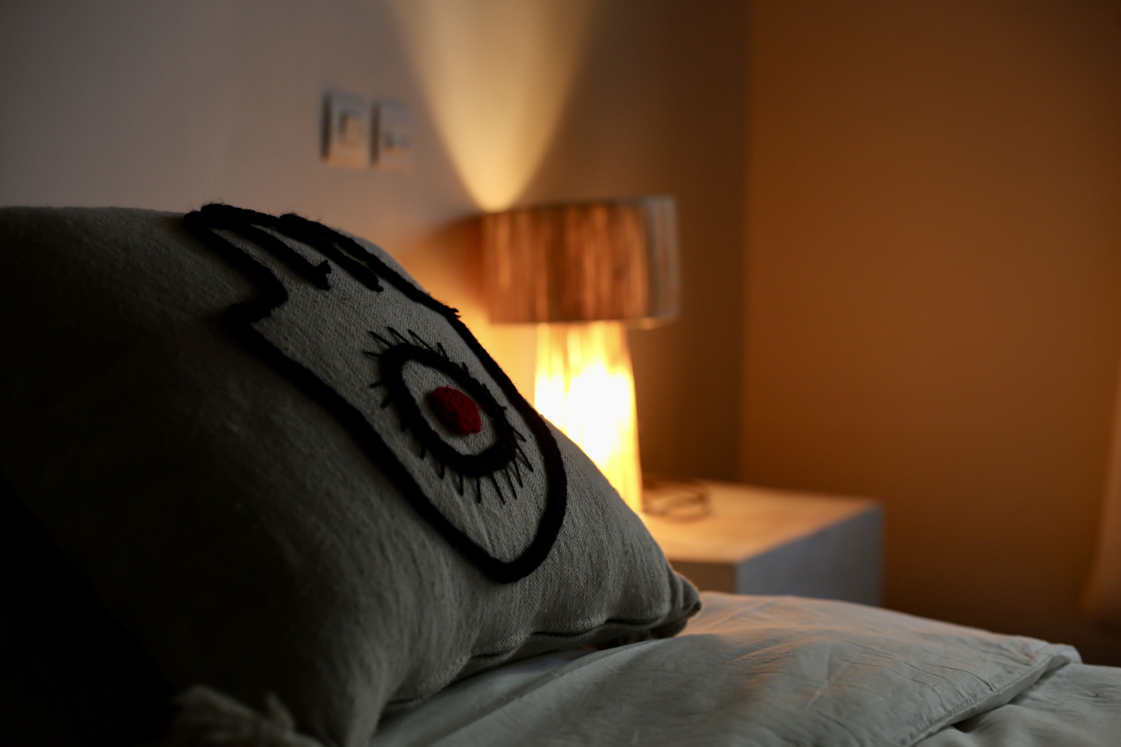 A bed with a pillow featuring a stitched eye design, a bedside lamp, and a small table with a lamp.