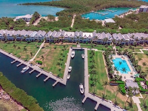 An aerial view of a resort with multiple bungalow-style buildings, a marina with boats, and a central swimming pool surrounded by palm trees.