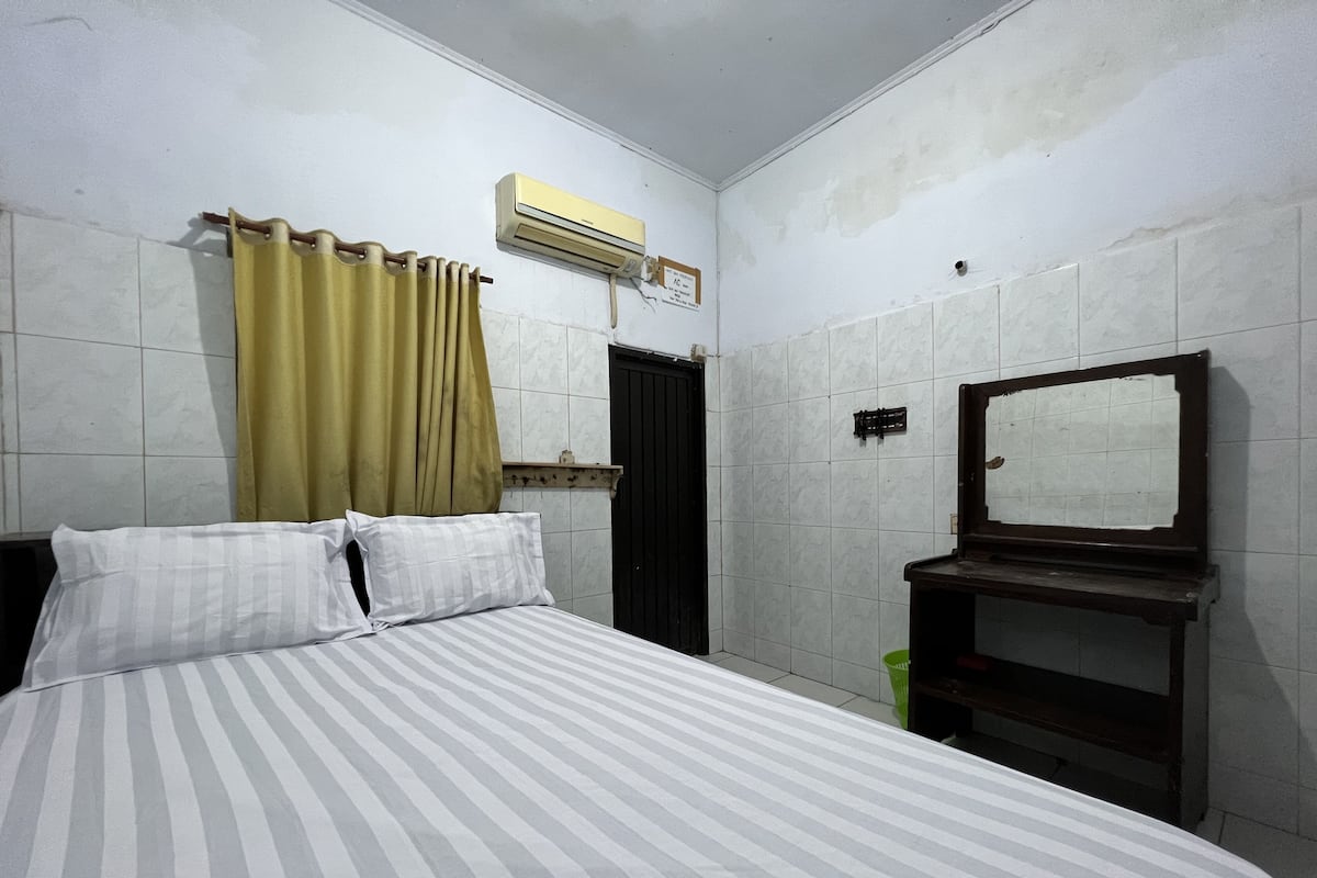 Une petite chambre d’hôtel simple, avec un lit simple, un miroir, une petite étagère et une fenêtre avec des rideaux.
