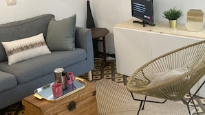 Una sala de estar con un sofá gris, una mesa de centro de madera con tazas rosas, una silla de mimbre, un escritorio pequeño con una computadora y pastos decorativos tipo pampa.