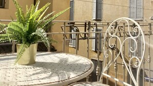 Un balcón con una mesa y sillas redondas de metal, una helechera en maceta y un edificio con balcones y ventanas al fondo.