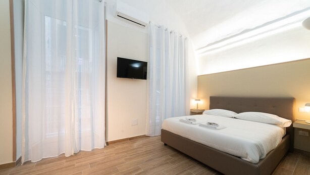 A hotel room with a bed, curtains, a flat-screen TV, and a nightstand with a lamp. - Villari 48 Apartment - Appartamento 2 (Naples)