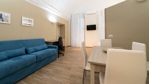 A modern living room with a blue sofa, a dining table with chairs, a TV mounted on the wall, and a window with curtains. - Villari 48 Apartment - Appartamento 2 (Naples)