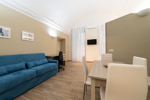 A modern living room with a blue sofa, a dining table with chairs, a TV mounted on the wall, and a window with curtains.