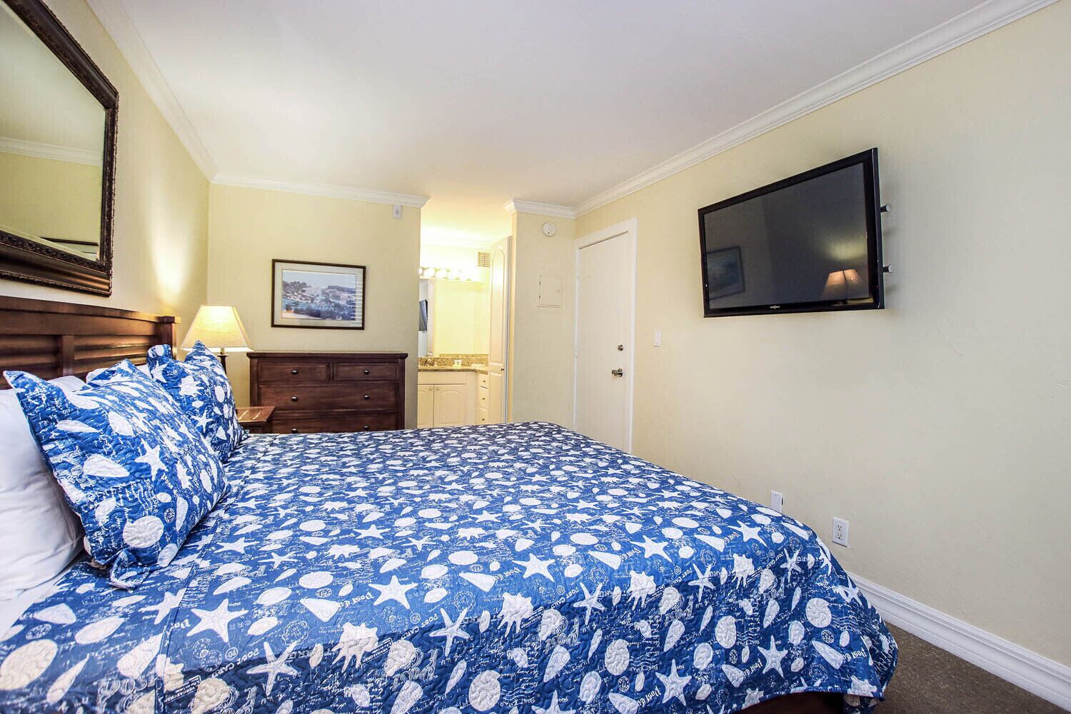 Une chambre à coucher comprenant un lit, une commode, un téléviseur fixé au mur et une porte donnant accès à une salle de bain.