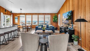 A spacious living room with a large window overlooking a beach, a blue sofa, a coffee table, and a TV mounted on the wall. - Stunning Home: Spacious layout, perfect for gathering with friends or family. (Rockaway Beach)