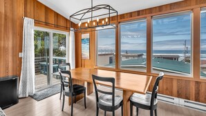 A dining area with a wooden table and chairs, large windows with a view of the ocean, and a ceiling light fixture. - Stunning Home: Spacious layout, perfect for gathering with friends or family. (Rockaway Beach)