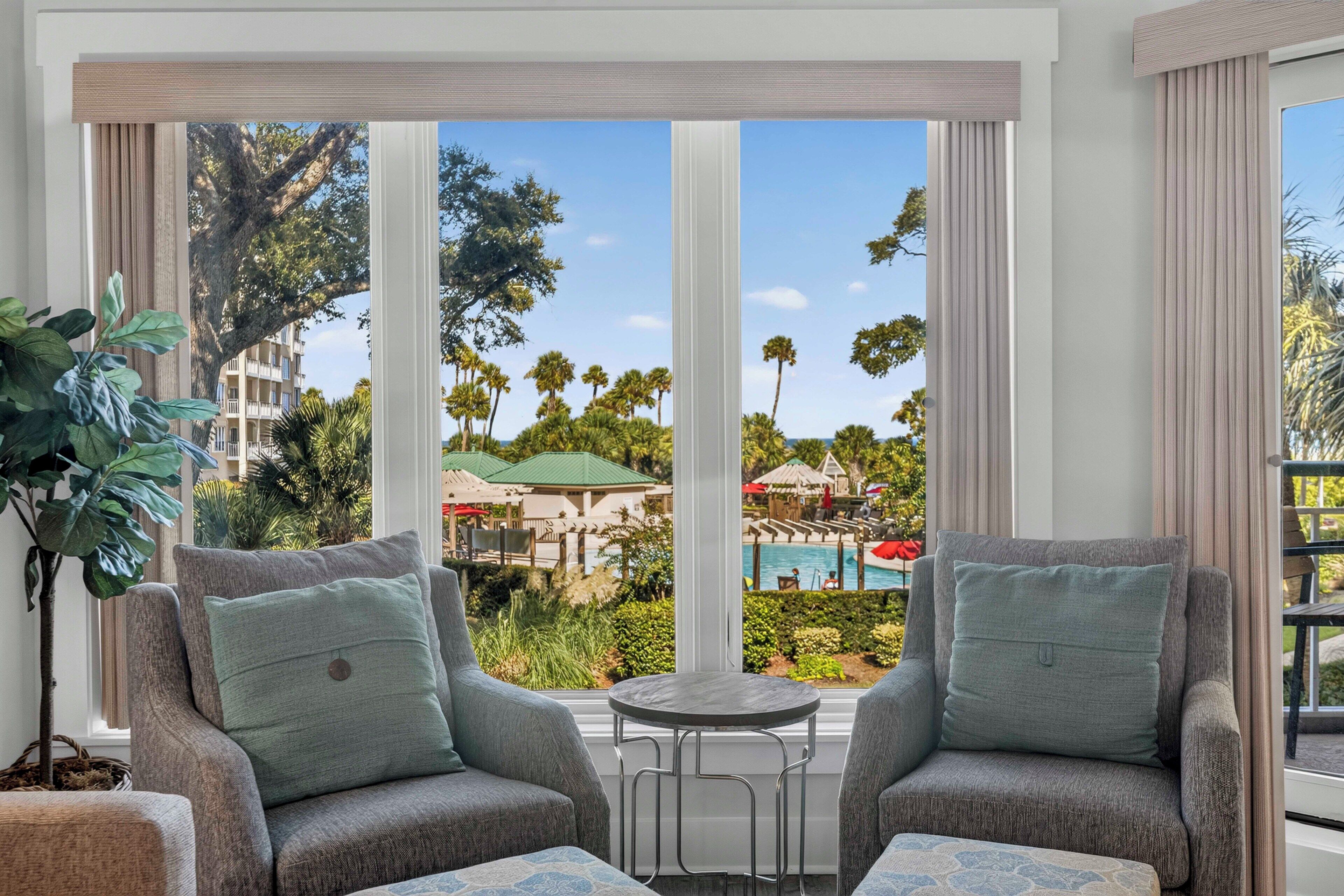 A room with two grey armchairs, a small round table, and a view of a pool and palm trees.