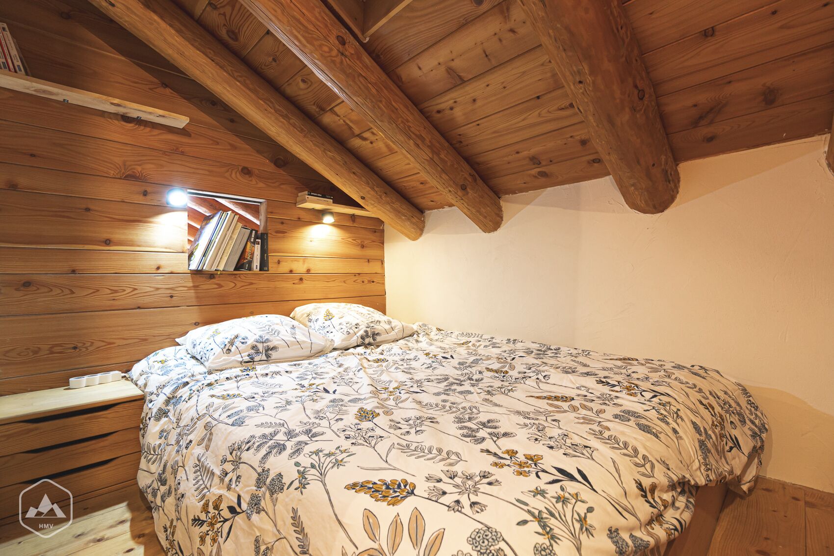 A bedroom with a wooden ceiling, a bed with floral bedding, a bedside table, and a shelf with books.