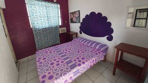 Un dormitorio con una cama, un ventilador de techo, una ventana con cortinas estampadas, un aire acondicionado de pared y una mesita.