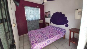 Un dormitorio con cama, ventilador de techo, ventana con cortinas estampadas, aire acondicionado de pared y una pequeña mesa de madera.