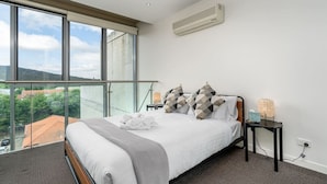 A modern hotel room with a large bed, a view of the city through floor-to-ceiling windows, and a balcony with a glass railing.