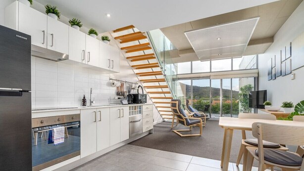 A modern kitchen with white cabinets, a stainless steel refrigerator, and a wooden staircase leading to a balcony.