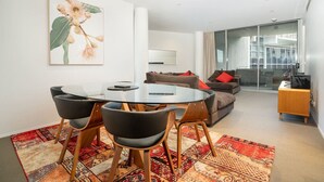 A modern living room with a round glass table, black chairs, a brown sofa with red cushions, and a large window offering a view of the city. - 2BR Apartment near Parliament House (Barton)