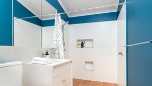 Et badeværelse med et hvidt toilet, en hvid vask med et håndklæde, en bruser med et geometrisk bruseforhæng og en hylde med toiletartikler.
