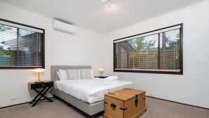 Een slaapkamer met een bed, nachtkastjes, een houten kist en grote ramen met jaloezieën.