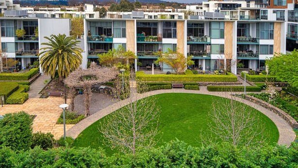 Un immeuble moderne à plusieurs étages, doté de balcons, d’une cour intérieure avec une pelouse et entouré de verdure.