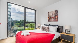 Een moderne hotelkamer met een groot raam, een bed met een rode deken, een nachtkastje met een lamp en een schilderij aan de muur.
