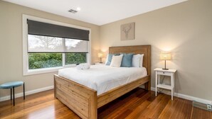 A bedroom with a wooden bed, a window with a dark roller blind, a bedside table with a lamp, and a wall-mounted artwork.