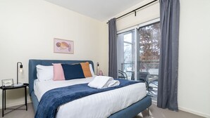 A bedroom with a bed, a nightstand, a window with curtains, and a view of the outside. - Captivating 3BR/2BA APT close to Dickson Shopping! (Lyneham)