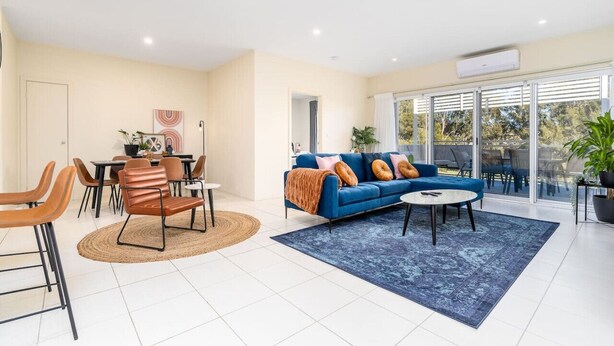 A modern living room with a blue sofa, orange chairs, a dining table, and a large window with a view of trees. - Captivating 3BR/2BA APT close to Dickson Shopping! (Lyneham)