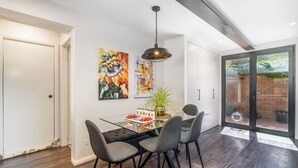 Un área de comedor con una mesa de vidrio, cuatro sillas, una lámpara colgante y dos cuadros en la pared.