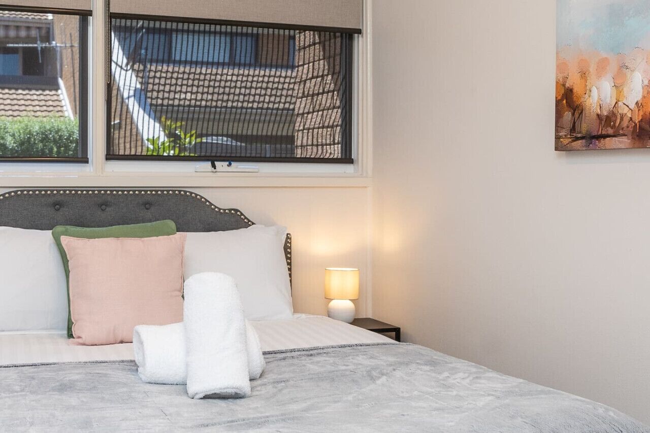 Un lit bien fait, avec des oreillers et une serviette enroulée, une lampe de chevet et une fenêtre donnant sur un immeuble.