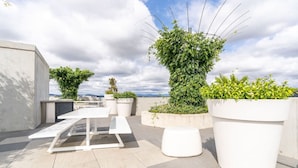 Une terrasse sur le toit, avec une table blanche et des bancs, entourée de grands pots de plantes et offrant une vue dégagée sur le ciel.