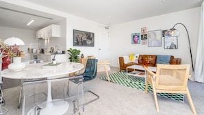 A modern living room with a round dining table, clear chairs, a sofa, and a coffee table. There are framed pictures on the wall and a lamp on a side table. - 2BR Penthouse w/ Breathtaking View! (Braddon)