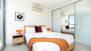 A modern hotel room with a large bed, a bedside table with a lamp, a framed picture on the wall, and a large mirror reflecting the room. - 2BR Penthouse w/ Breathtaking View! (Braddon)