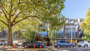 A modern building with a glass facade, parked cars, and a tree in the foreground. - 2BR Penthouse w/ Breathtaking View! (Braddon)