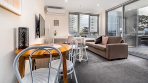 A modern living room with a wooden dining table, white chairs, a beige sofa, and a large window offering a city view. - 2BR Apt in the heart of the Gungahlin Town Centre! (Gungahlin)