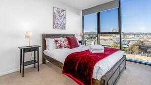 Een slaapkamer met een groot raam, een bed met witte beddengoed, een rode plaid en een klein nachtkastje met een lamp.