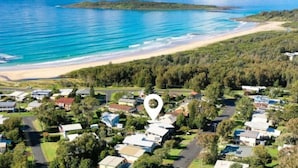 An aerial view of a coastal residential area with houses, a beach, and a forested island in the distance. - Lavish Home 4mins to the beach Pet- Friendly House (Bawley Point)