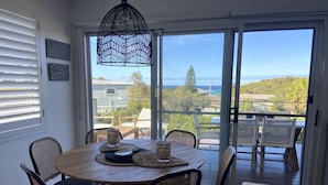 A dining area with a round table and chairs, a hanging light fixture, and a view through a glass door to an outdoor deck with chairs. - Lavish Home 4mins to the beach Pet- Friendly House (Bawley Point)