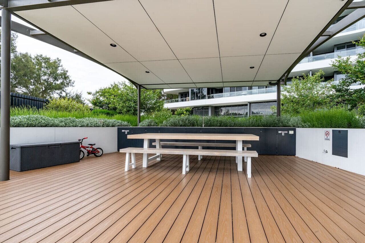 An outdoor wooden deck with a bench and a bicycle, surrounded by greenery and a modern building in the background.