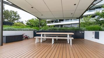An outdoor wooden deck with a bench and a bicycle, surrounded by greenery and a modern building in the background.