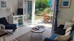 A living room with a sofa, a coffee table, a TV on a cabinet, and a sliding glass door to a patio with outdoor furniture. - Appartement Accueillant Tout Proche mer (Clohars-Carnoët)