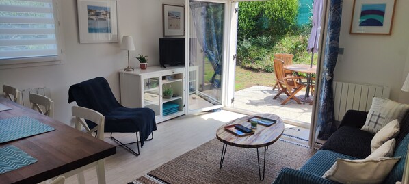 Un salon comprenant un canapé, une table basse, un téléviseur posé sur un meuble et une porte-fenêtre coulissante donnant sur une terrasse aménagée avec du mobilier d’extérieur.