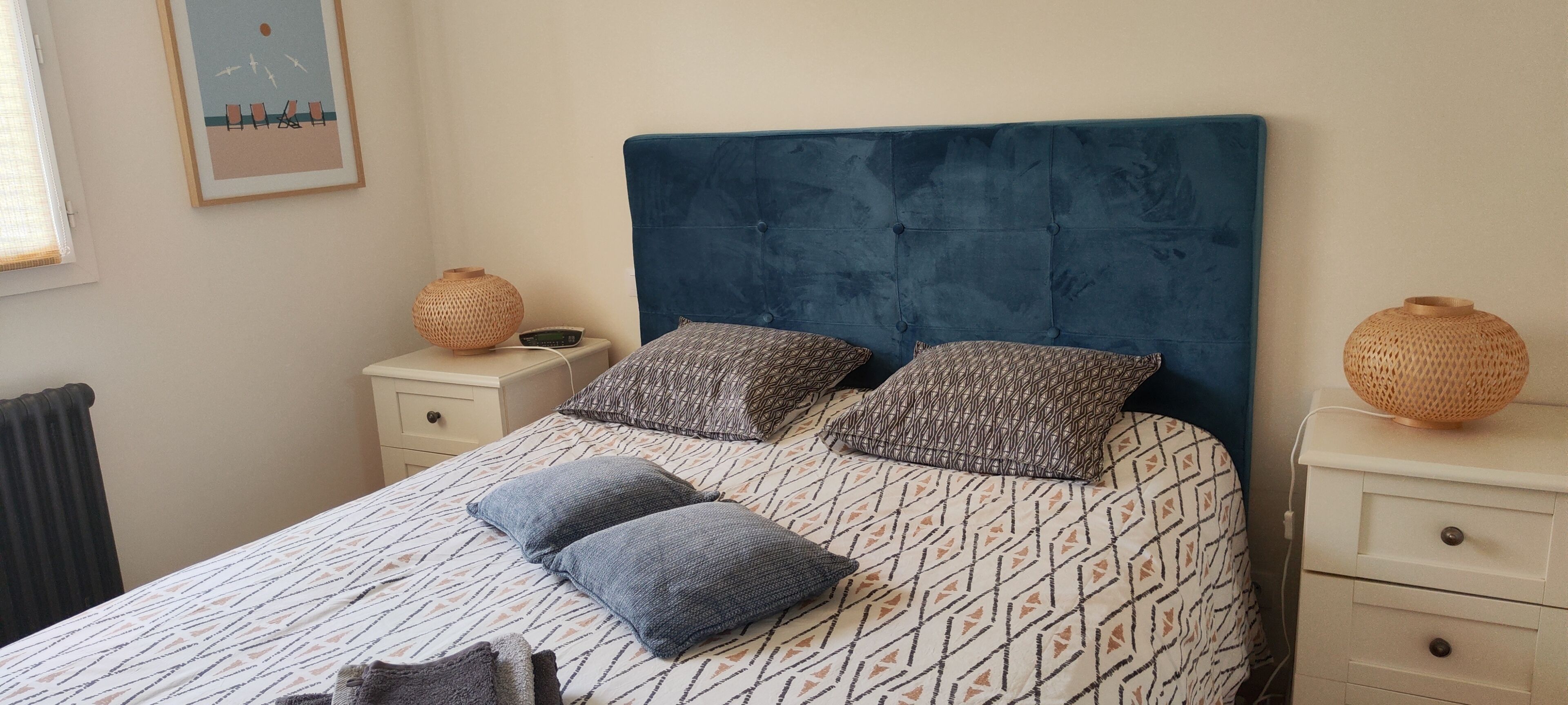 A neatly made bed with patterned bedding and two pillows, a blue upholstered headboard, and two bedside tables with woven lamps.