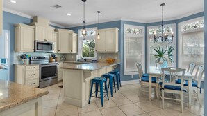 A kitchen with white cabinets, a central island with blue barstools, a dining table with chairs, and a chandelier. - Spacious 3BR, Dreamy Backyard, 2 Pools, 6-Min Stroll to Beach + Free Tickets! (Santa Rosa Beach)