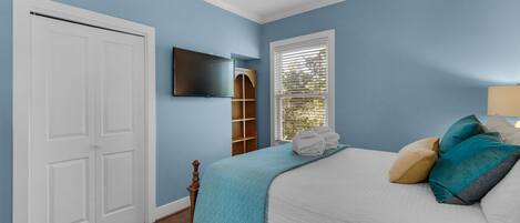 A bedroom with a bed, a TV, a window with blinds, and a door.