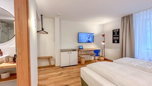 Une chambre d’hôtel avec un plancher en bois, un lit, un bureau avec une chaise, une télévision et une fenêtre avec des rideaux.