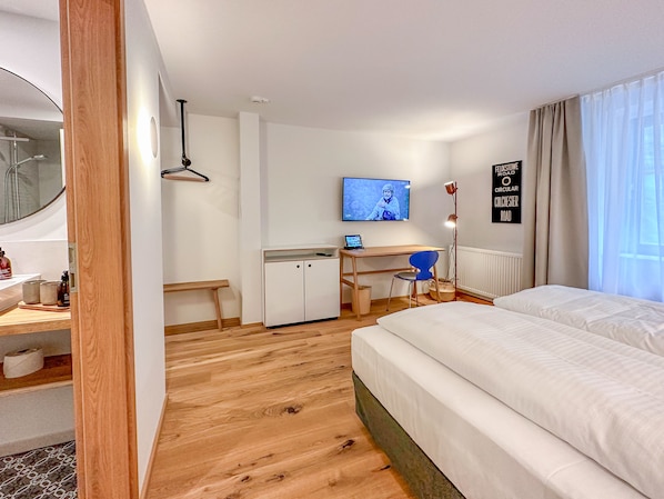 A hotel room with a wooden floor, a bed, a desk with a chair, a TV, and a window with curtains.