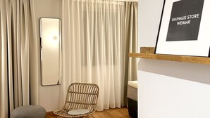 Une pièce avec un grand miroir, un fauteuil en osier, un pouf gris et un panneau encadré accroché au mur.