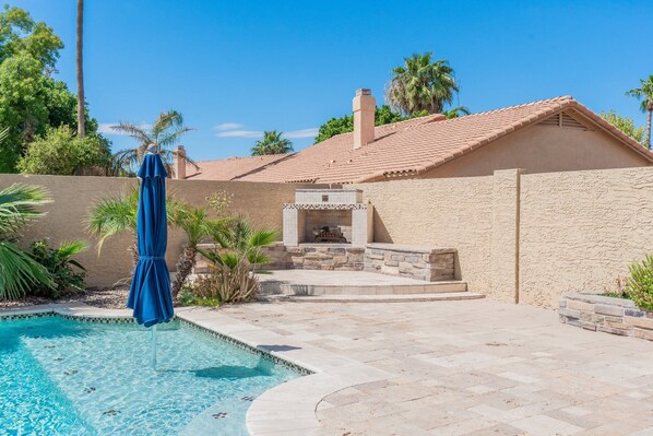 A swimming pool with a blue umbrella, a stone fireplace, and a stucco building with a tiled roof.