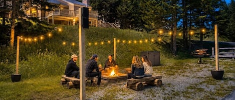 En gruppe mennesker sidder omkring et bål i et naturområde med træer og en bygning i baggrunden.
