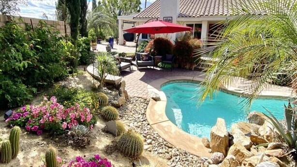 A pool area with a red umbrella, surrounded by cacti and desert plants, and a house with a tiled roof in the background. - Private pool, spa, waterfall, firepit, BBQ, tennis, gym, hiking trails, casino (Palm Desert)