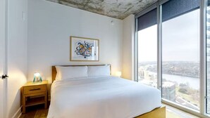 A modern bedroom with a large bed, a nightstand with a lamp, a framed abstract painting, and a large window offering a city view.