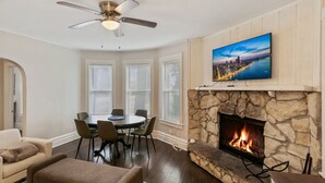 Una sala de estar con chimenea de piedra, un televisor montado encima y un comedor con mesa redonda y sillas.